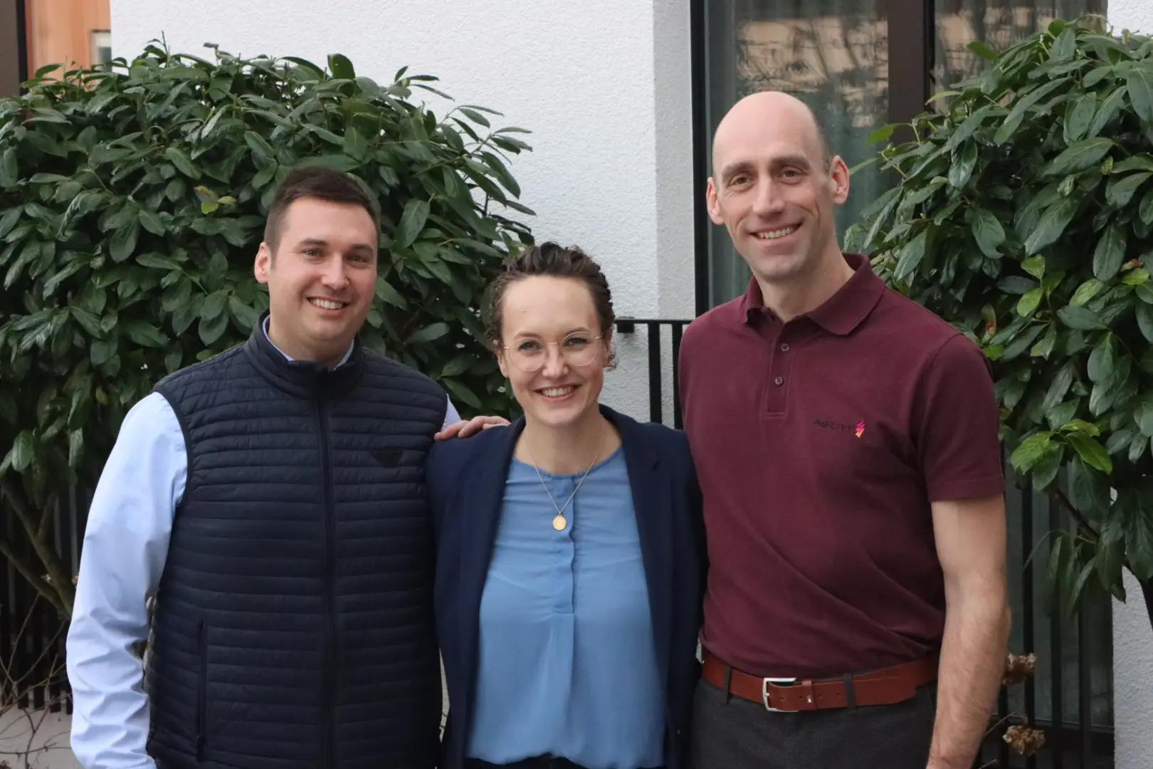 Ulrich Zahner in dunkelblauer Weste und hellem Hemd, Theresa Gerster in blauem Top und dunkelblauem Blazer und Rainer Bickel in bordeaux-farbenem Polo, Geschäftsleitung Allgeier inovar, stehen nebeneinander vor Hauswand und Kirschlorbeer-Gebüsch.