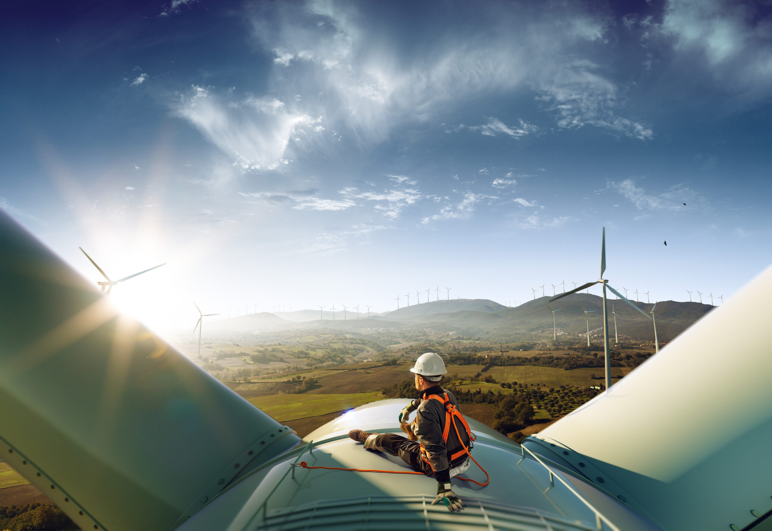 Monteur sitzt auf Windkraftanlage und schaut in die Ferne, wo in einer Hügellandschaft weitere Windkraftanlagen stehen betrieben von AURELO Energiepark Manager von Allgeier inovar.