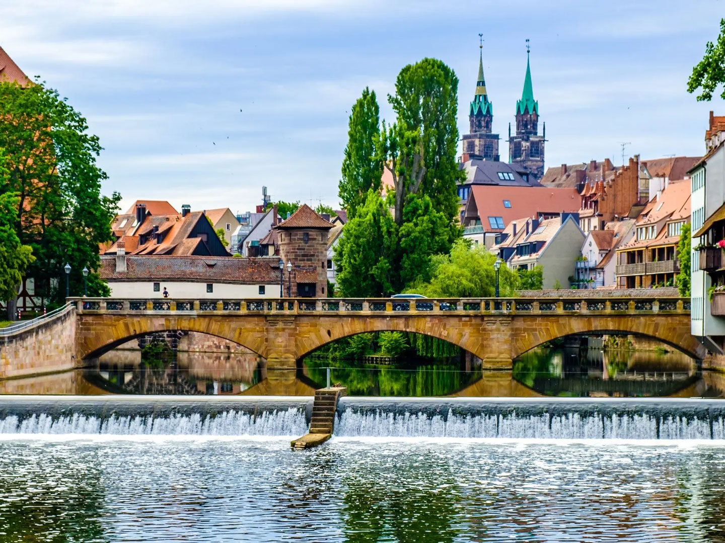 Allgeier Inovar mit Standort in Nürnberg, zu erkennen an der Pfeilerbrücke über die Pegnis im Vordergrund und den Kirchtürmen im Hintergrund. Dazwischen grüne Bäume und Häuser vor blauem Himmel.