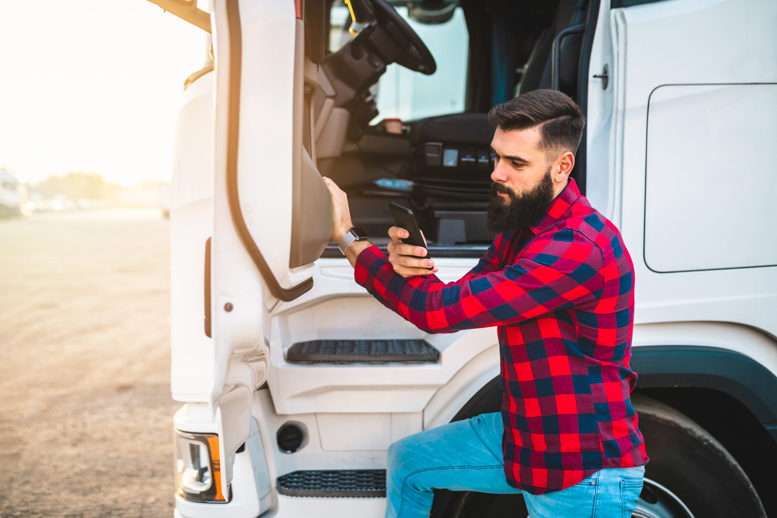 Mann mit schwarzem Bart und rotkariertem Hemd steht vor Trucktür und schaut auf Handy mit der Software ServiceApp von Allgeier inovar.