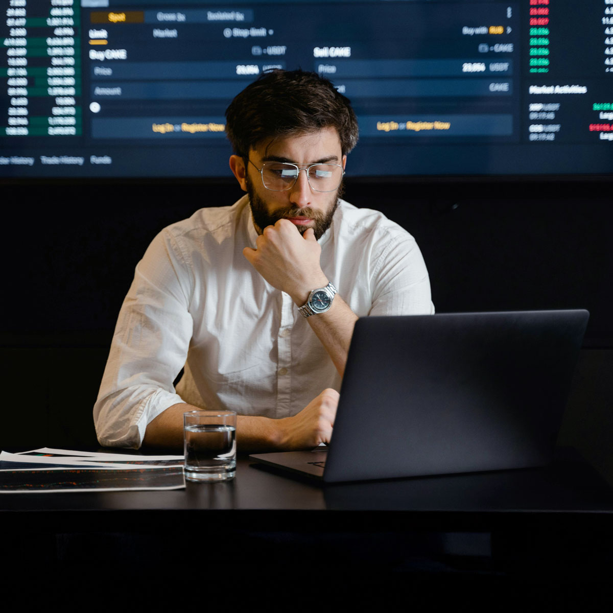 Mann mit dunklem Haarschopf, Bart und Brille sitzt am Tisch und schaut in einen Laptop mit Software von Allgeier inovar. Daneben ein Glas Wasser, im Hintergrund ein Datenmonitor.