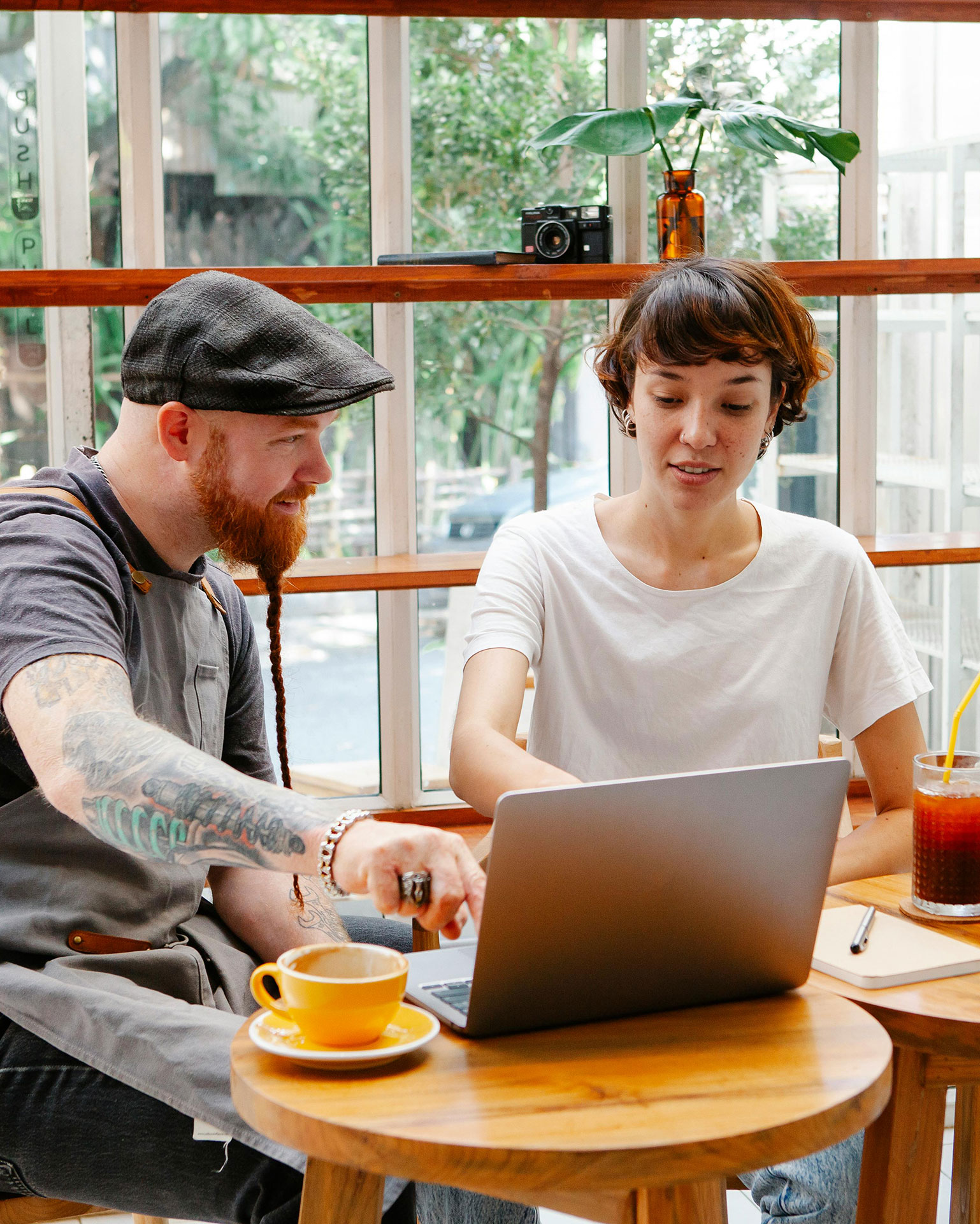 Mann mit Kappe, rotem Bart und Tatoo sitzt am Tisch und bespricht mit Frau in weißem T-Shirt vor einem Laptop die Buchhaltungslösung syntona e finac von Allgeier inovar.