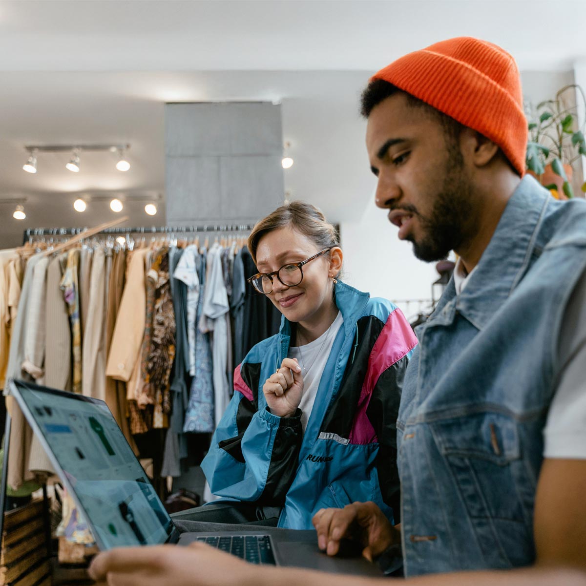 Mann mit Brille und Ballonseide-Jacke und Mann mit Jeansweste und oranger Mütze schauen in eine Laptop mit der Webshop-Lösung Allgeier Commerce von Allgeier inovar. Im Hintergrund ein Kleiderständer.