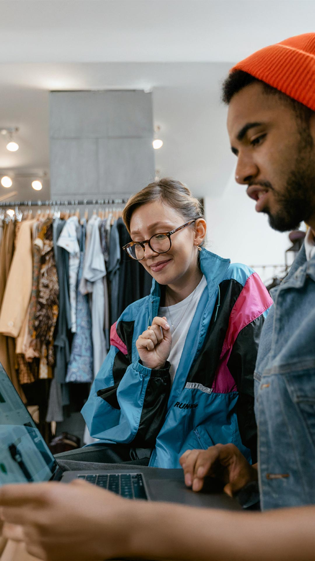 Mann mit Brille und Ballonseide-Jacke und Mann mit Jeansweste und oranger Mütze schauen in eine Laptop mit der Webshop-Lösung Allgeier Commerce von Allgeier inovar. Im Hintergrund ein Kleiderständer.