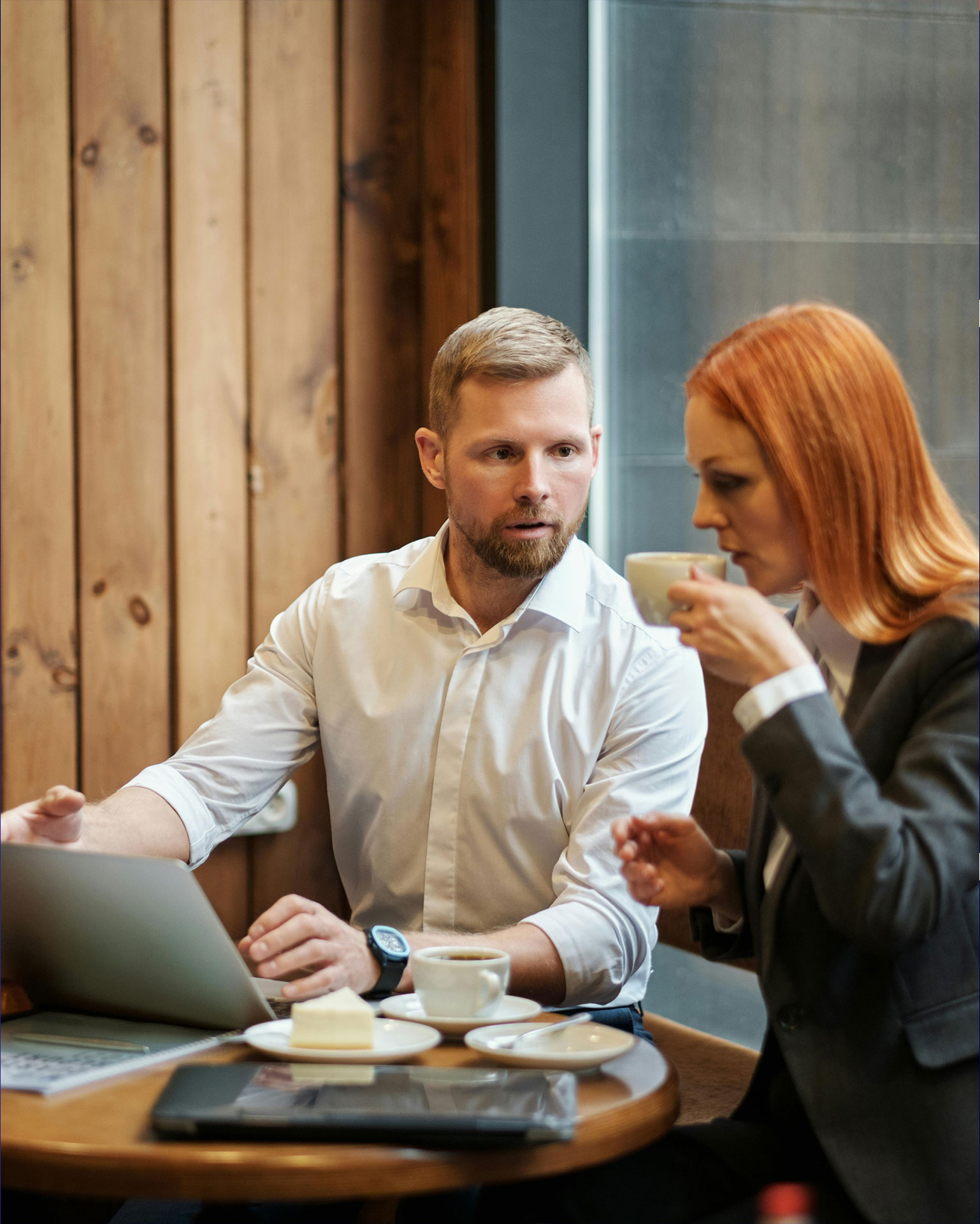 Mann mit weißem Hemd und Bart vor Laptop an Bistotisch schaut zu Frau mit roten Haaren, die eine Kaffeetasse zum Mund führt und bespricht die Lösung für E-Rechnungen von Allgeier inovar.