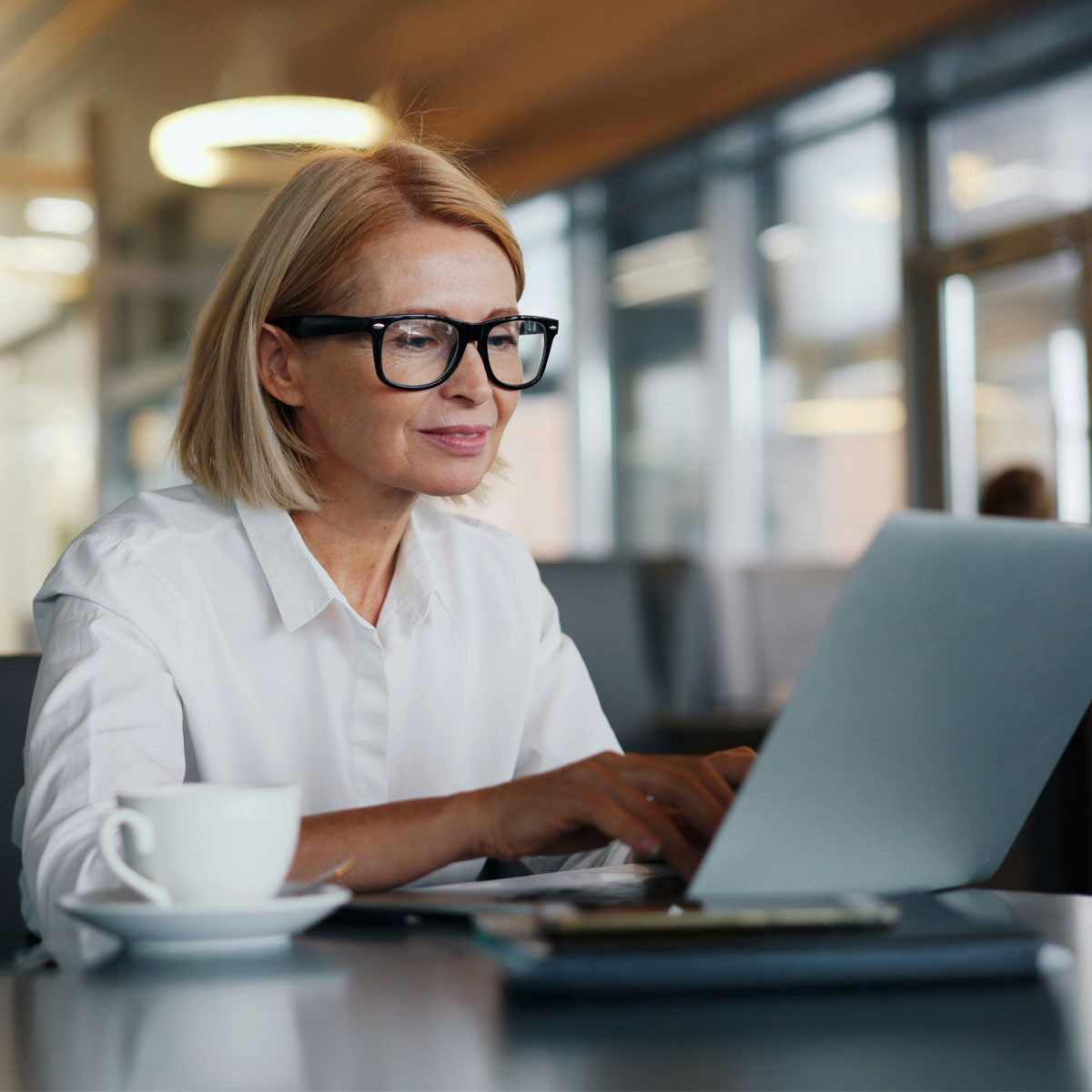 Blonde Frau mit schwarzer Brille in weißer Bluse sitzt mit Kaffeetasse am Tisch vor Laptop und beschäftigt sich mit der Software von Allgeier inovar für gesetzliche Änderungen.