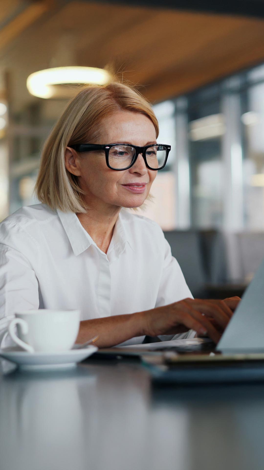 Blonde Frau mit schwarzer Brille in weißer Bluse sitzt mit Kaffeetasse am Tisch vor Laptop und beschäftigt sich mit der Software von Allgeier inovar für gesetzliche Änderungen.