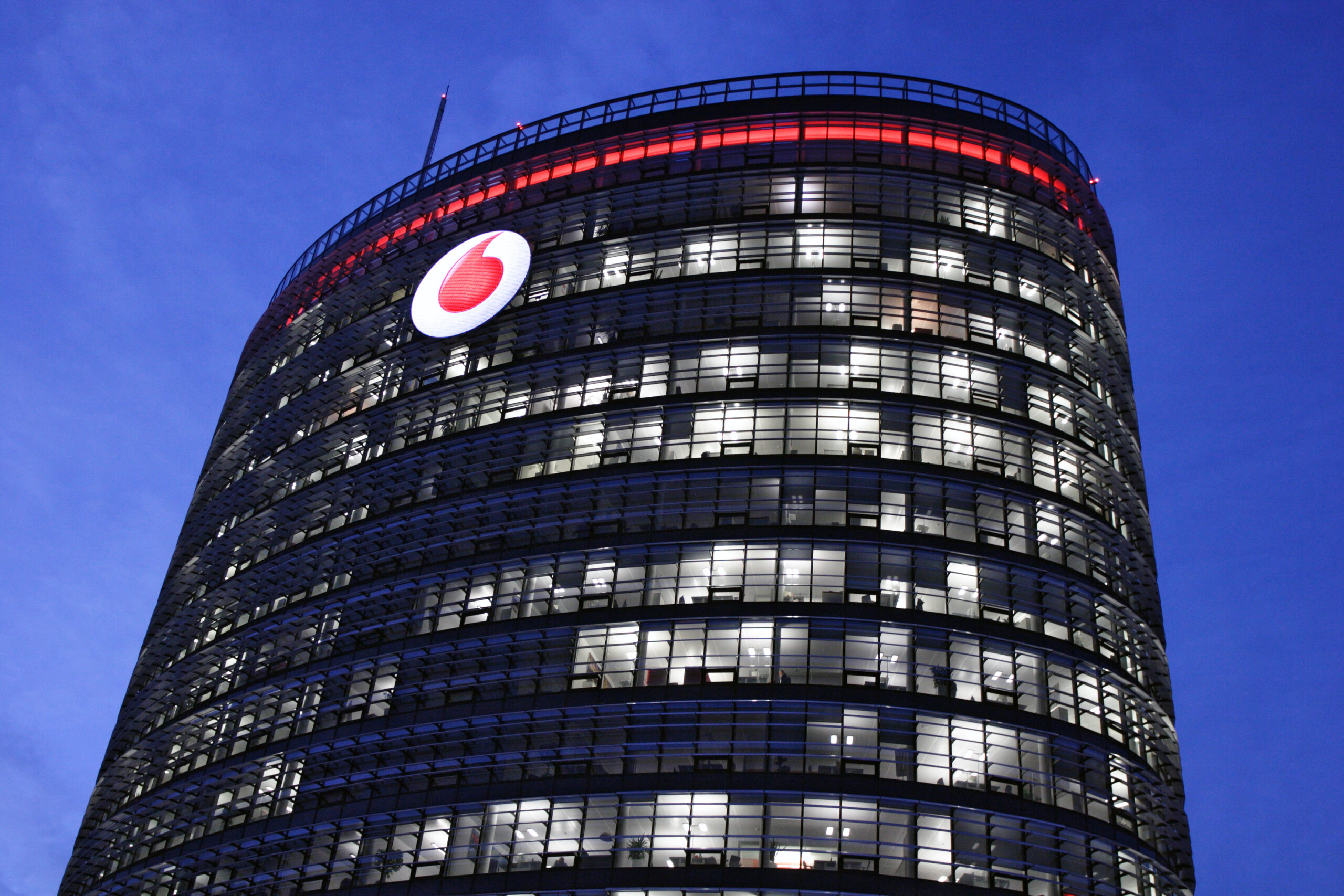 Vodafone-Hochhaus mit Logo in der Dämmerung mit erleuchteten Fenstern, die mit metasonic® von Allgeier inovar die Rechnungen steuern.