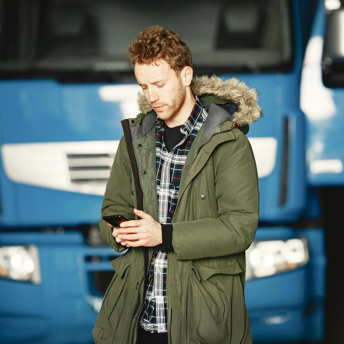 Mann in grünerm Parka und kariertem Hemd steht vor blauem LKW und arbeitet mit Handy auf der die ServiceApp von Allgeier inovar installiert ist.