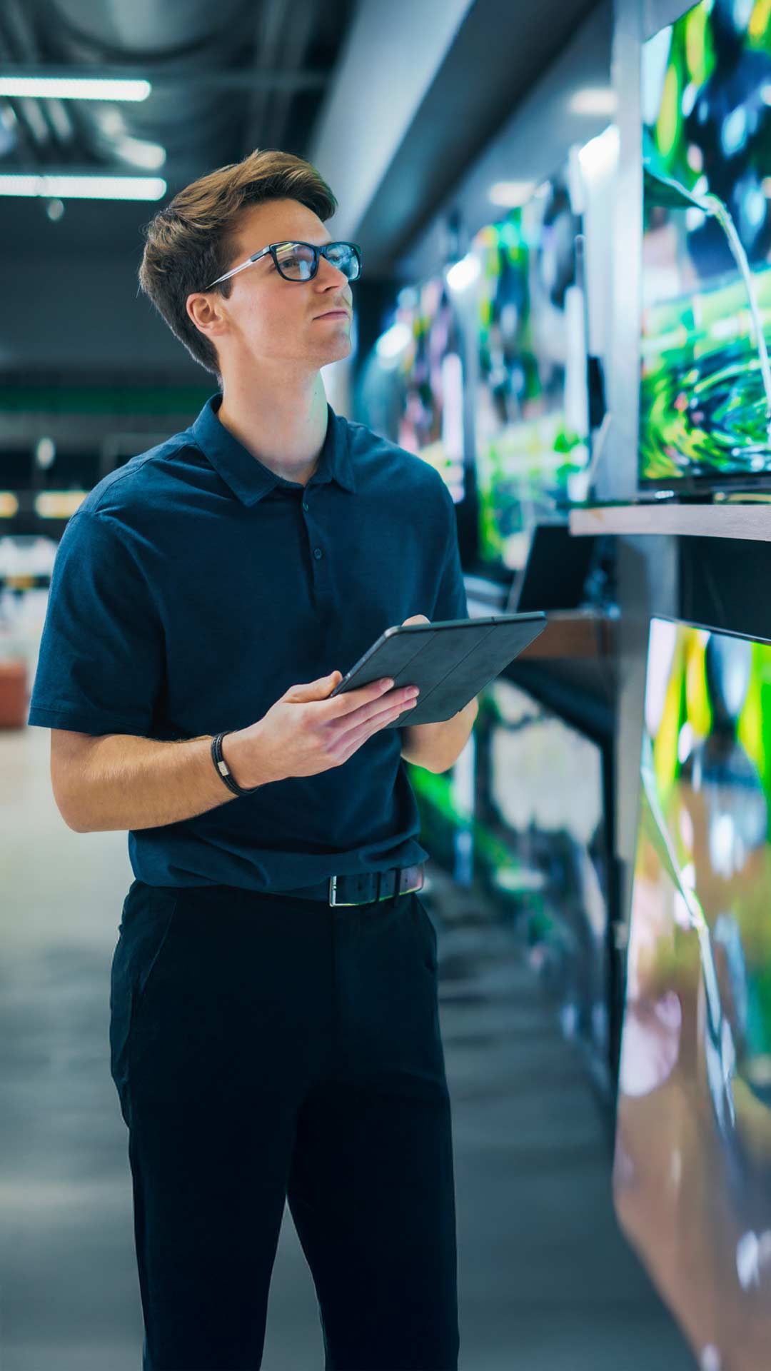 Mann mit Tablet in der Hand und schwarzer Brille und blauem Polo schaut auf Monitore und Großhandel und organisiert das Ordermanagement mit der OrderApp von Allgeier inovar.