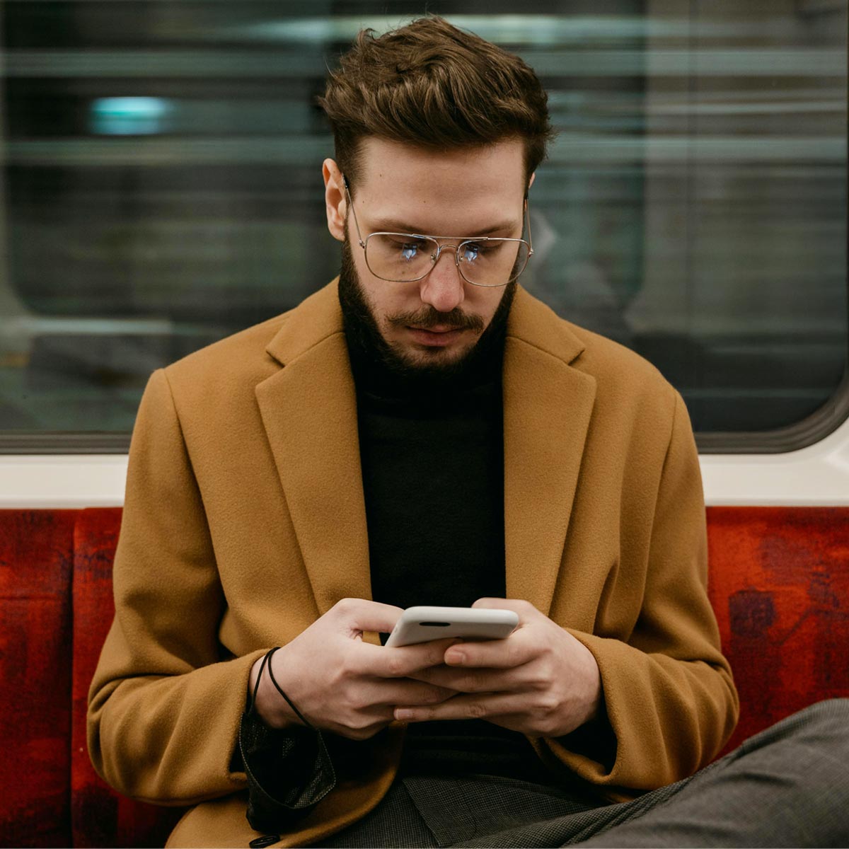 Mann mit dunklem Rolkragne und braunem Mantel sitzt in S-Bahn mit Handy und schaut auf die syntona® logic App von Allgeier inovar.