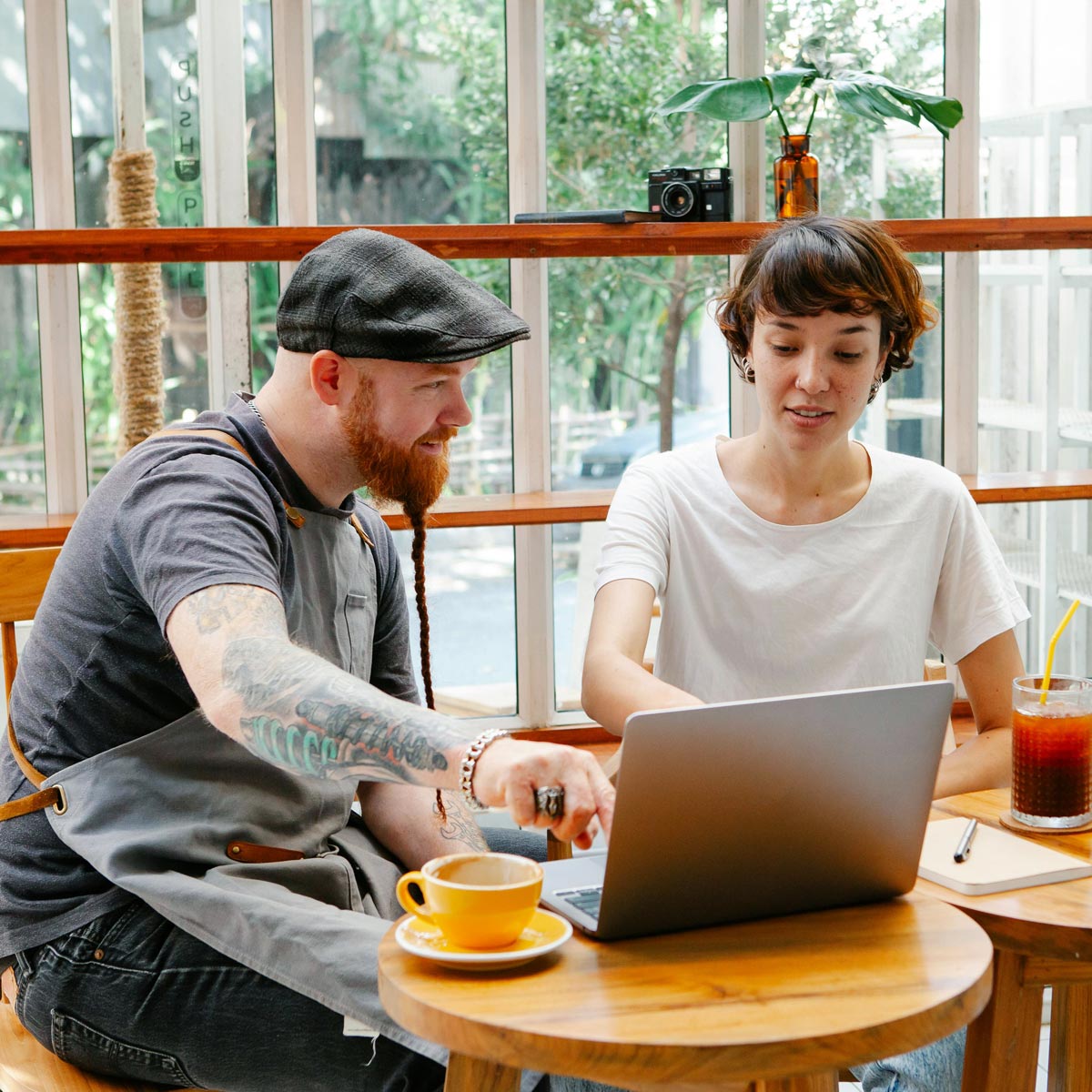 Mann mit Kappe, rotem Bart und Tatoo sitzt am Tisch und bespricht mit Frau in weißem T-Shirt vor einem Laptop die Buchhaltungslösung syntona e finac von Allgeier inovar.