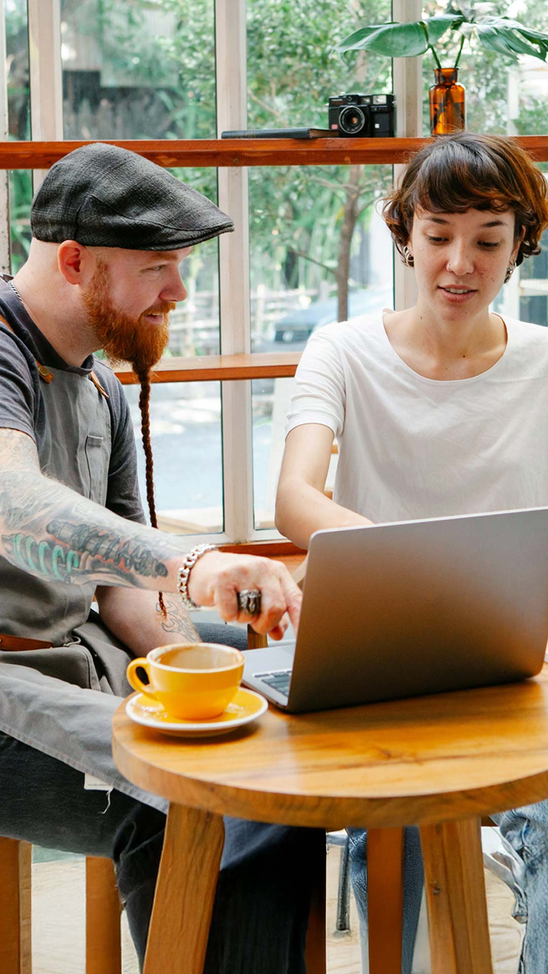 Mann mit Kappe, rotem Bart und Tatoo sitzt am Tisch und bespricht mit Frau in weißem T-Shirt vor einem Laptop die Buchhaltungslösung syntona e finac von Allgeier inovar.