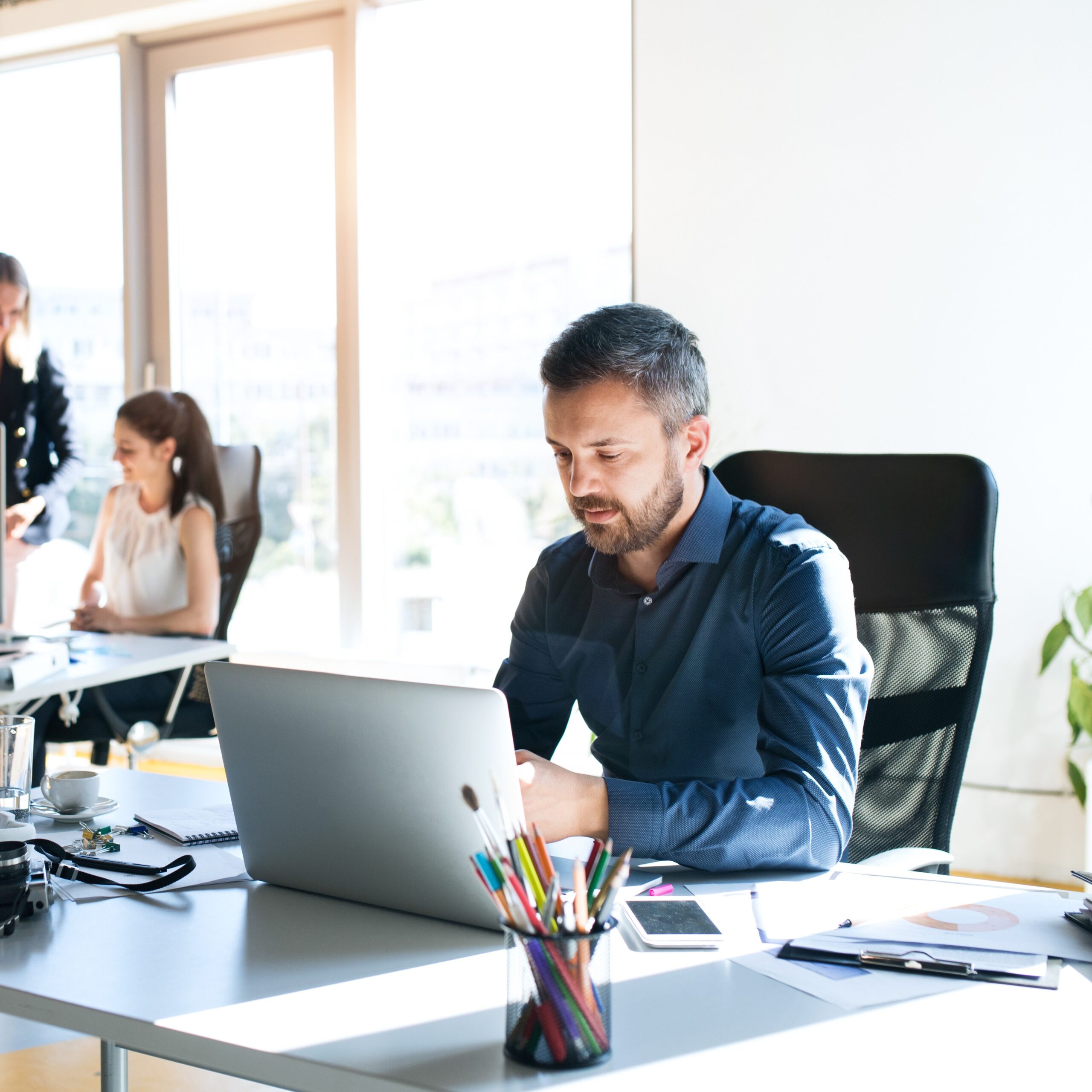 Mann sitzt vor Laptop im Büro, im Hintergrund stehen zwei Frauen sprechend am Computer