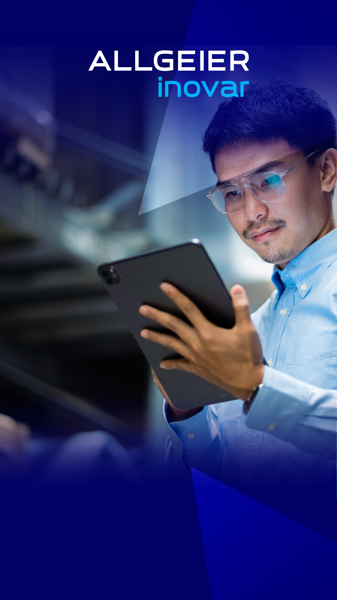 Ein Mann steht in einem Lager und hat ein Tablet in der Hand. Er trägt ein hellblaues Hemd und eine Brille und hat dunkle schwarze Haare.